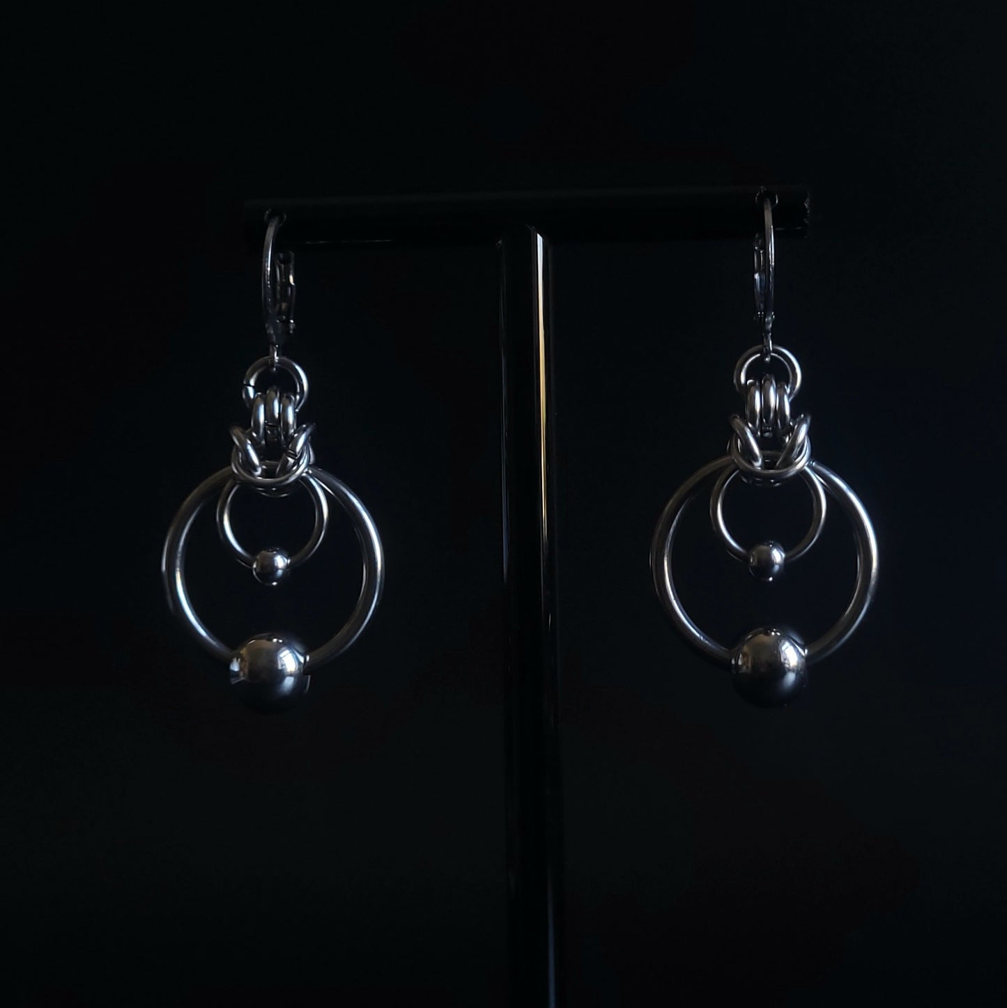 Face-on view of ORBIT Earrings with silver huggie hoops, interlocking rings, polished stainless steel ball charms, and half Byzantine chainmail weave, showcasing chunky O ring earrings for gothic and alternative jewelry style.